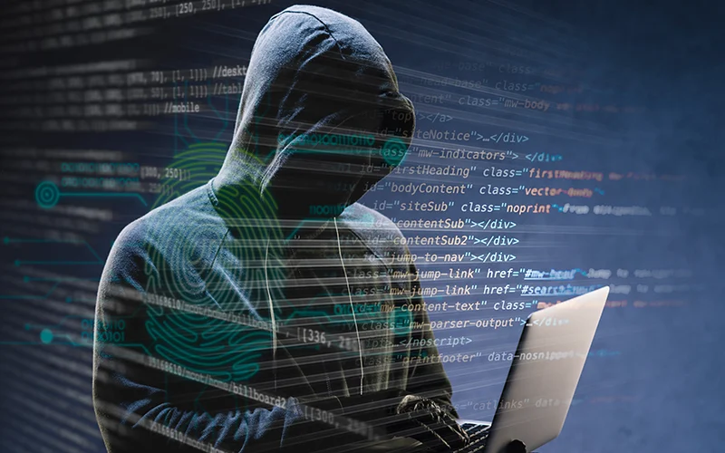 A hooded figure using a laptop, representing how cyber attacks happen with malicious hackers exploiting security vulnerabilities.